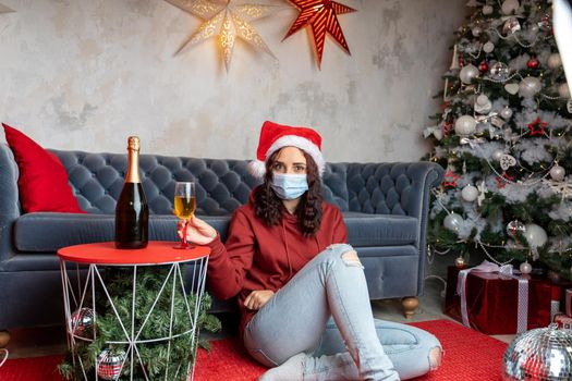 Young woman in medical mask and Santa Claus hat sitting on floor near sofa in room. Alone charming brunette in protective mask celebrating of safe Christmas during coronavirus pandemic