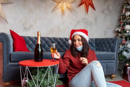 Young woman in medical mask on beard and santa hat takes glass of champagne, sitting on floor near sofa in room. Charming brunette smiles, celebrating of safe Christmas during coronavirus pandemic