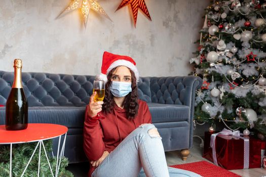 Young woman in medical mask and santa hat with glass of champagne sits on floor near sofa in room. Charming brunette in protective mask celebrating of safe Christmas during coronavirus pandemic