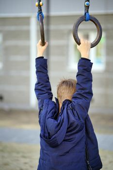 Little child swings on sports rings in yard of house. Active boy playing on playground