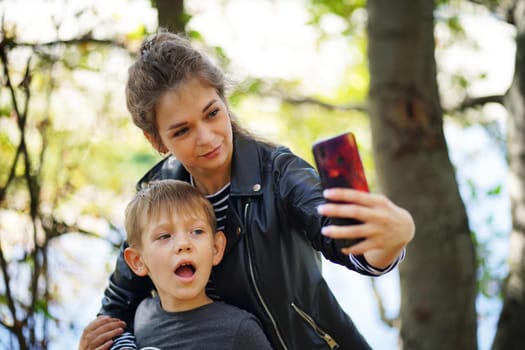 A young mother with her son takes a selfie. Smiling young woman with long hair and a child take a selfie with a smartphone, in the park.