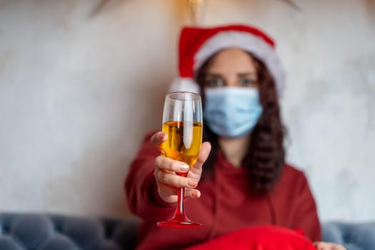 Close up of young woman in medical mask holds glass of champagne. Brunette in protective mask offers toast, celebrating of safe Christmas during coronavirus pandemic