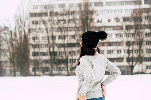 Rear view of young woman in gray knitted sweater and hat standing on street in winter season. Female looking at high-rise building in cloudy weather