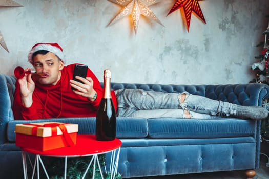 Young handsome man in Santa Claus hat with glass of champagne taking selfie on smartphone. Funny guy photographing himself on mobile phone and lying on couch. Concept of Christmas celebration at home