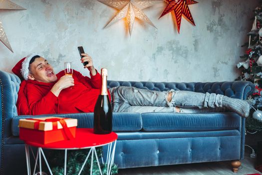 Young man in Santa Claus hat browses smartphone and drinks champagne, lying on couch. Handsome guy resting with mobile phone and alcohol. Concept of Christmas celebration at home