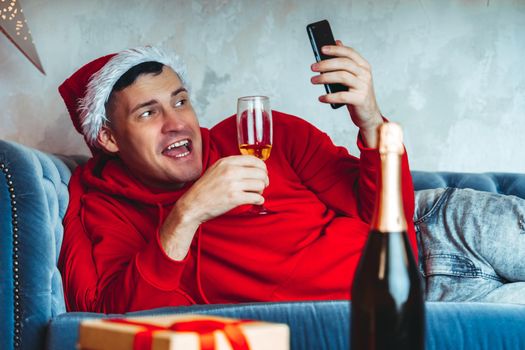 Young handsome man in Santa Claus hat with glass of champagne conducting live broadcast with friends on smartphone. Happy guy celebrating Christmas online on internet and lying on couch at home