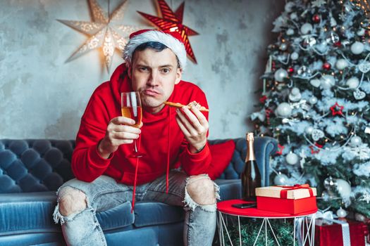 Young man in santa hat drinks champagne and eats pizza, sitting on sofa in room. Happy male resting with food and alcohol. Concept of Christmas celebration at home