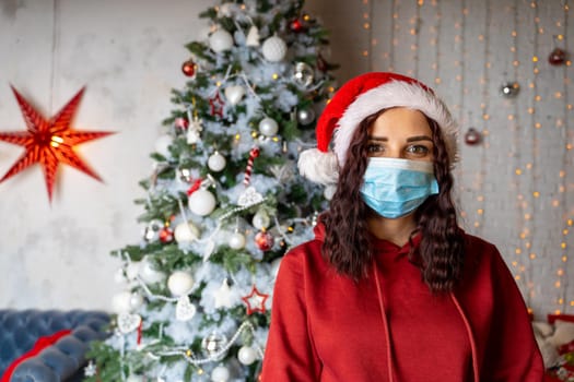 Young woman in medical mask and santa hat on background of Christmas tree. Charming brunette in protective mask celebrating of safe Christmas during coronavirus pandemic