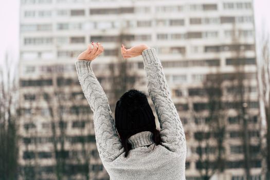 Rear view of young woman in gray knitted sweater and hat standing on street in winter season. Female looking at high-rise building and stretching her hands in cloudy weather