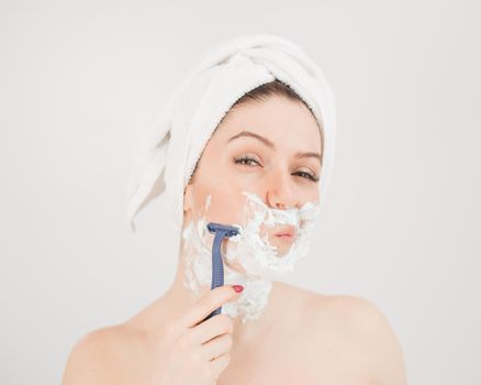 Cheerful caucasian woman with a towel on her head and shaving foam on her face holds a razor on a white background.