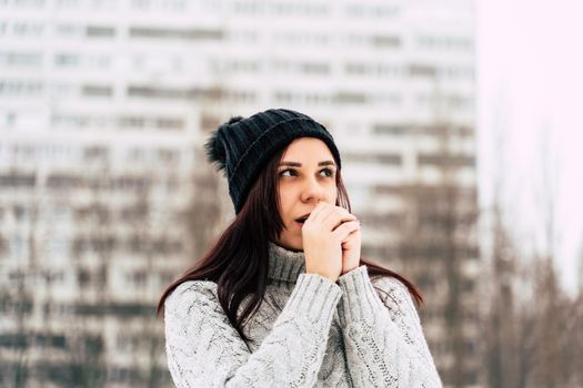 Portrait of young woman in gray knitted sweater and hat on background of high-rise building. Close up of pretty female warming hands with her breath on walk in winter season