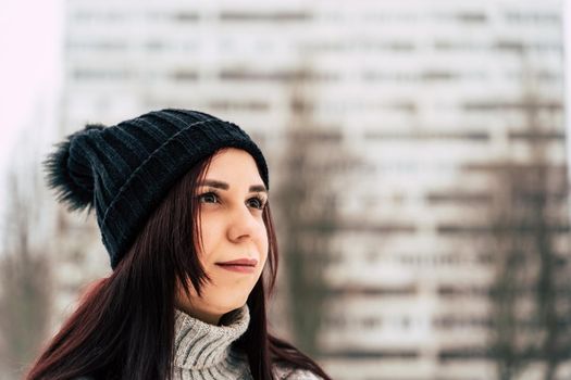 Thoughtful young woman in gray knitted sweater and hat standing on street in winter season. Pretty brunette looking away and reflecting about something in cloudy weather