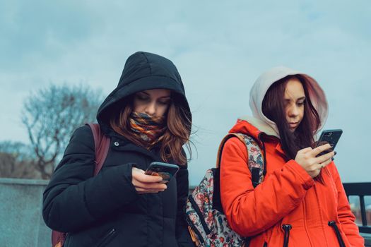 Close up of young women browsing smartphone in city park. Pretty females in hoods using mobile phone