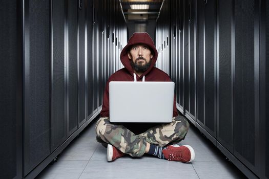 The hacker in a hood sits with the computer among servers, the burglar. Storage of data, cloudy services, system breaking, attack of hackers. High quality photo