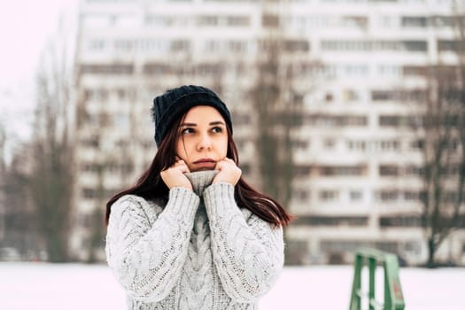 A pretty girl, dressed in a gray knitted sweater and a hat, covers her face with a sweater collar.