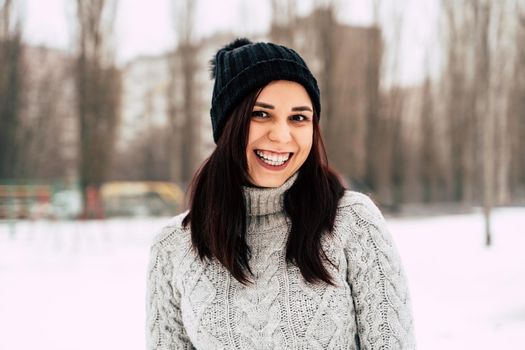 Young, beautiful woman with winter cap and gray sweater smiling and laughing while standing outside in winter.