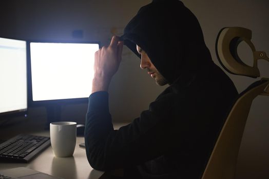 Computer hacker. Young man in black hoodie using multiple computers for stealing data while sitting in dark room. Binary code. Cyber attack. Cyber security