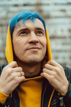 Portrait of young man in hood on background of high-rise building. Handsome guy with blue hair posing on city street in springtime