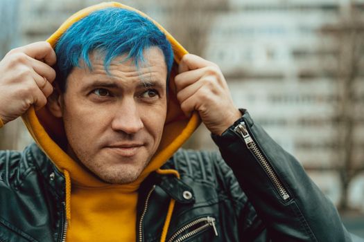 Portrait of young man in hood on background of high-rise building. Handsome guy with blue hair posing on city street in springtime