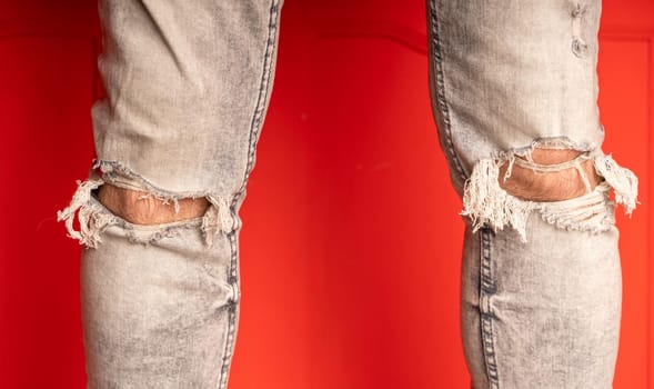 Close up of men's legs in jeans with holes on red background. Fashionable holes in grey jeans on unrecognizable man