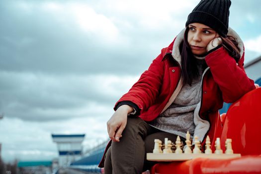 Young woman in winter clothes plays chess, sitting on stadium bleachers alone. Female in black cap with chess on sports stadium in cloudy weather