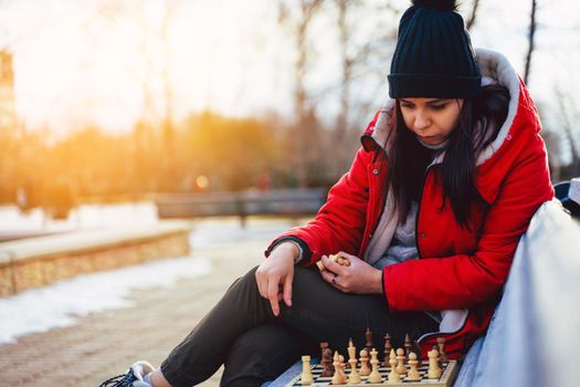 Young woman in winter clothes plays chess, sitting on bench in city park. Female in black cap with chess on wooden pew in cloudy weather