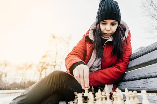 Young woman in winter clothes plays chess, sitting on bench in city park. Female in black cap with chess on wooden pew in cloudy weather