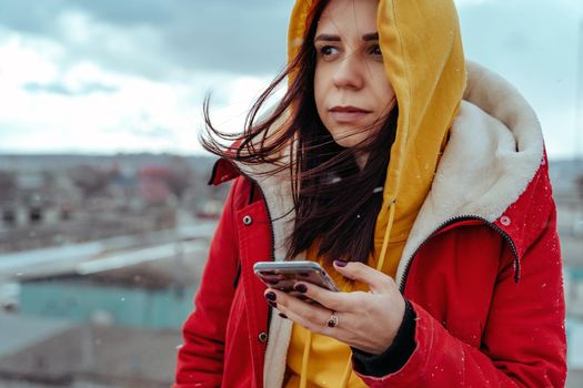 Portrait of young woman with mobile phone on roof of building. Brunette in yellow hoodie and red jacket browsing smartphone in cloudy weather