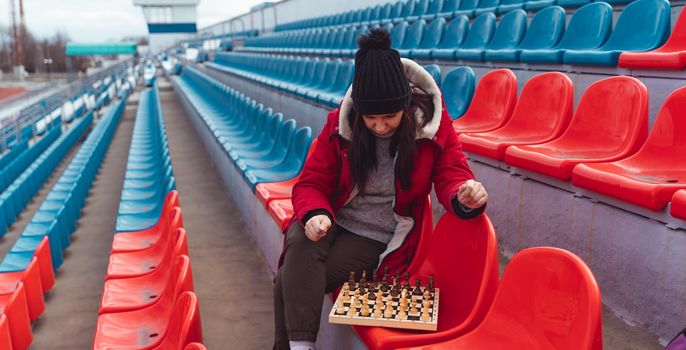 Young woman in winter clothes plays chess, sitting on stadium bleachers alone. Female in black cap with chess on sports stadium in cloudy weather