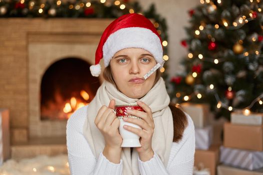 Upset female wearing santa hat, scarf and sweater, measuring temperature and drinking hot tea, holding cup with two hands, sitting near fireplace and x-mas tree.