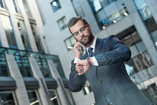 A young businessman with glasses and a beard negotiates a meeting. He has a smartphone in his hands and looks at his watch.