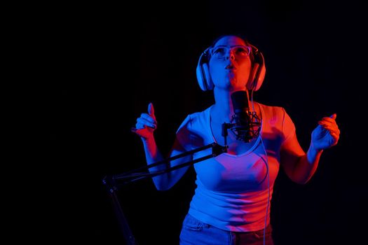 Caucasian woman in glasses and headphones sings into a microphone in neon light on a black background. An emotional girl is recording a song in a recording studio.