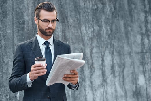 Young businessman in suit and glasses holding a paper cup and reading business newspaper in his hands