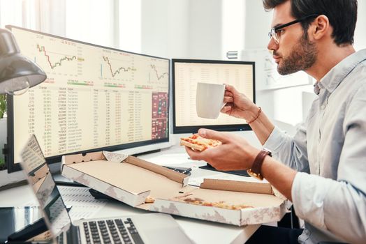 Food for productive work. Side view of young bearded trader in eyewear is eating hot pizza while looking at monitor screen with trading charts and financial data in his modern office. Coffee break. Food concept. Business concept. Trade concept