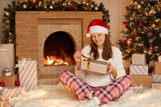 Caucasian woman opening present box while sitting on soft carpet in festive living room, lady wearing checkered pants, white shirt and santa claus hat, celebrating Christmas at home.