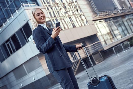 Young woman with suitcase smiling at her phone outside
