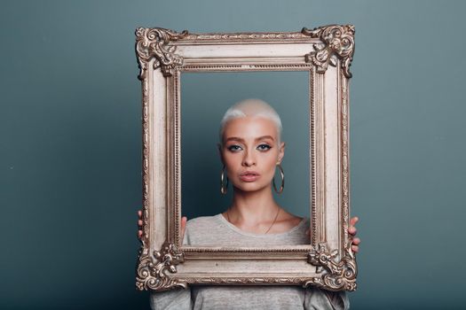 Millenial young woman with short blonde hair holds gilded picture frame in hands behind her face portrait