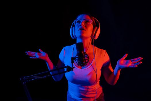 Caucasian woman in glasses and headphones sings into a microphone in neon light on a black background. An emotional girl is recording a song in a recording studio.