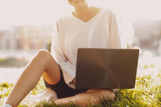 Portrait of young smiling millenial european short haired woman using laptop at green grass meadow in park. Beautiful happy blonde girl outdoor. Summer fashion female clothing.