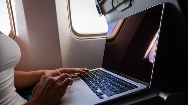 Faceless woman in a mask works on a laptop on an airplane while sitting by the window during the flight. Freelancer on a journey