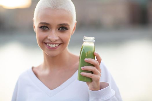 Portrait of young smiling millenial european short haired woman with green smoothie. Beautiful happy blonde girl outdoor.