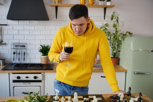 Young man playing chess on kitchen table and drinking red wine. Male with alcohol plays in logical board game with himself, standing in kitchen