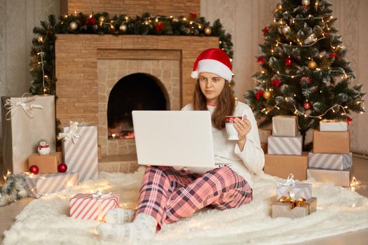 Serious woman having video call via lap top, talking to somebody or working online, Christmas holidays at home, lady wearing white casual sweater, checkered pants and santa hat, drinks hot tea.