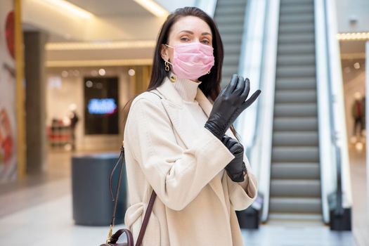 beautiful woman in pink medical mask putting on leather gloves in a public place on background escalator