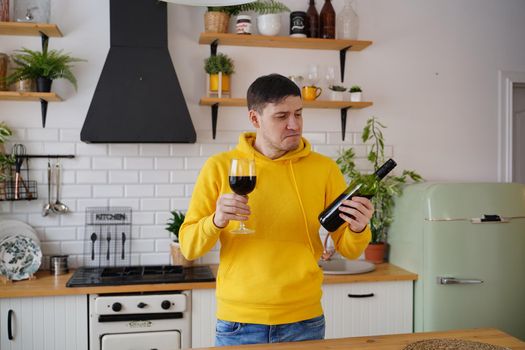 Relaxed young man drinks red wine and looks on bottle. Adult guy resting with alcohol in kitchen