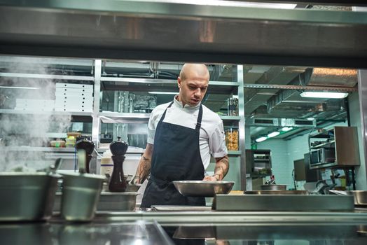 I should be careful. Serious male chef with several tattoos on his arms holding a frying pan above the oven in a restaurant kitchen. Cooking process