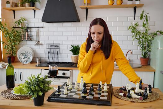 Young woman playing chess on kitchen table. Female plays in logical board game with herself in kitchen