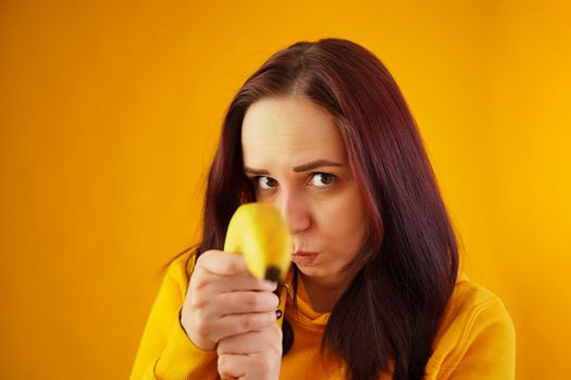 Portrait of young woman with banana on yellow background. Close up of female in yellow hoodie plays with fruit, imagining it as weapon