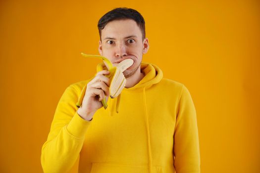 Portrait of young man eating banana on yellow background. Funny guy with bulging eyes in yellow hoodie shoved fruit in mouth