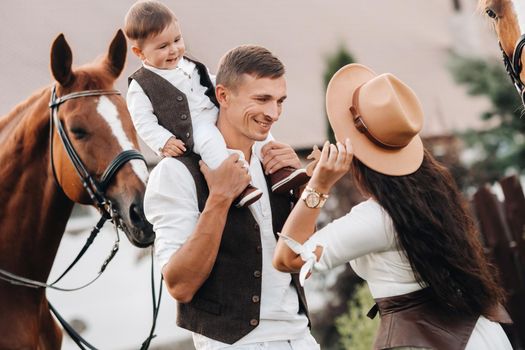 A family in white clothes with their son stand near two beautiful horses in nature. A stylish couple with a child are photographed with horses.